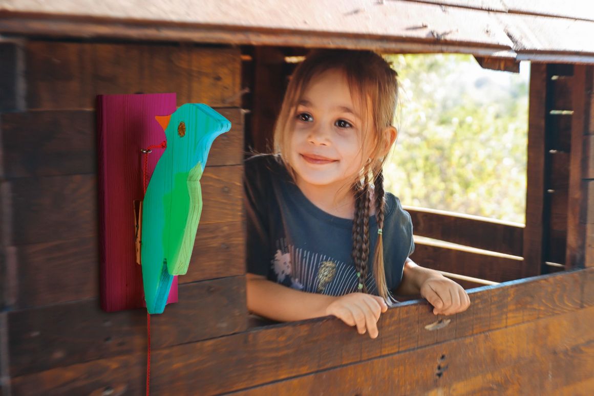 Kind in einem Spielhaus aus Holz mit buntem Holzvogel.